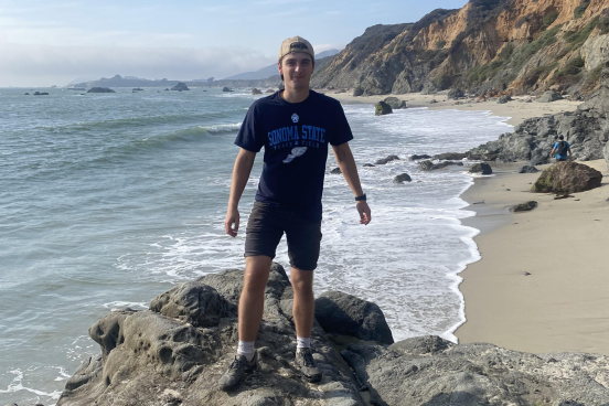 David is standing on a rock overlooking the ocean and beach. He is wearing a Sonoma State t-shirt and baseball hat.
