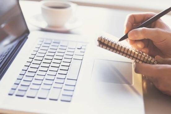 A person is writing in a notebook while a laptop sits on the table
