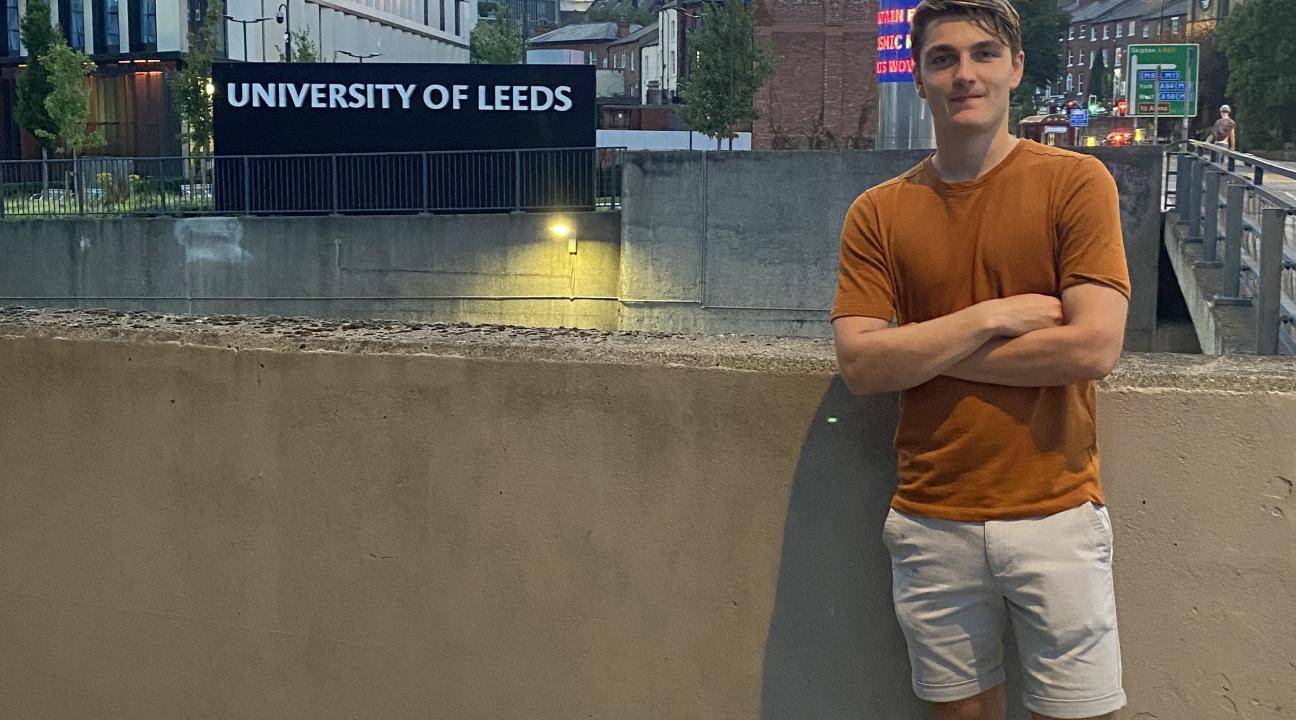 David is standing in front of a cement wall, behind him is a sign that reads 'University of Leeds' and the cityscape of Leeds
