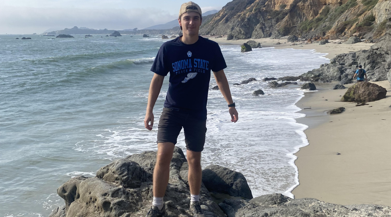 David is standing on a rock overlooking the ocean and beach. He is wearing a Sonoma State t-shirt and baseball hat.