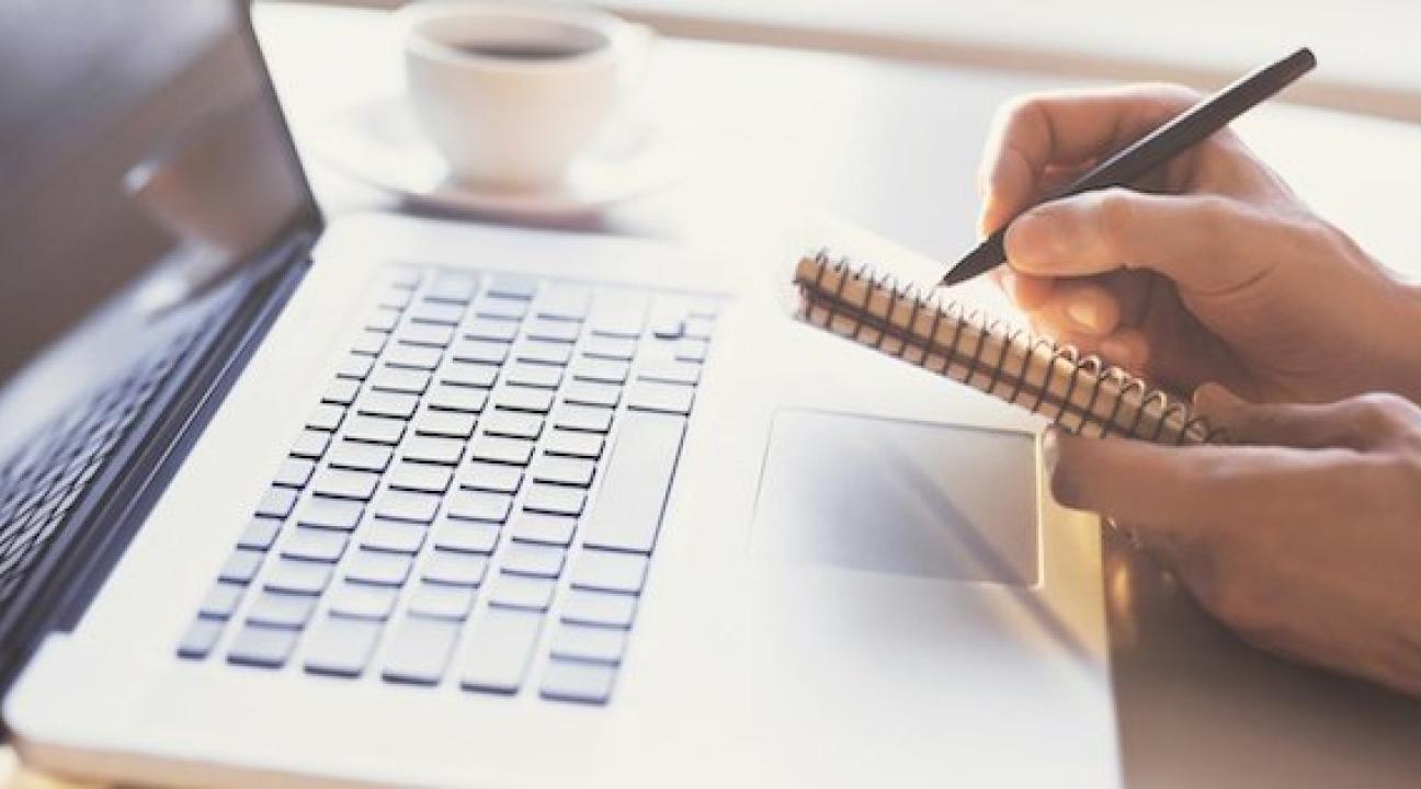 A person is writing in a notebook while a laptop sits on the table