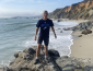 David is standing on a rock overlooking the ocean and beach. He is wearing a Sonoma State t-shirt an