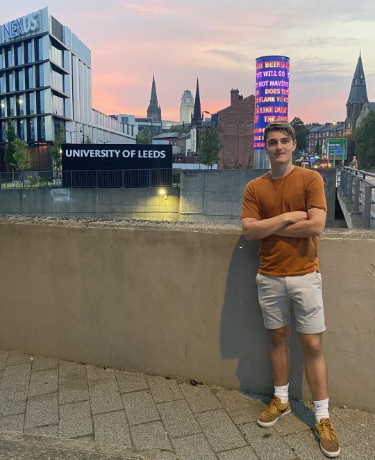 David is standing in front of a cement wall, behind him is a sign that reads 'University of Leeds' and the cityscape of Leeds