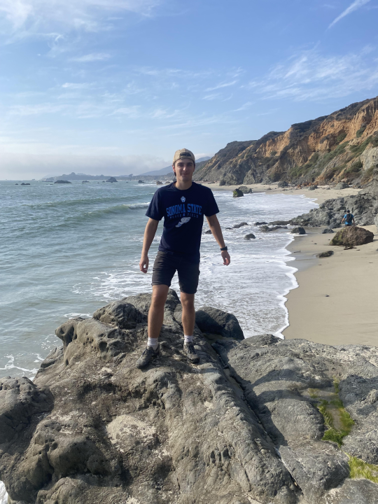 David is standing on a rock overlooking the ocean and beach. He is wearing a Sonoma State t-shirt and baseball hat.