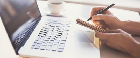 A person is writing in a notebook while a laptop sits on the table